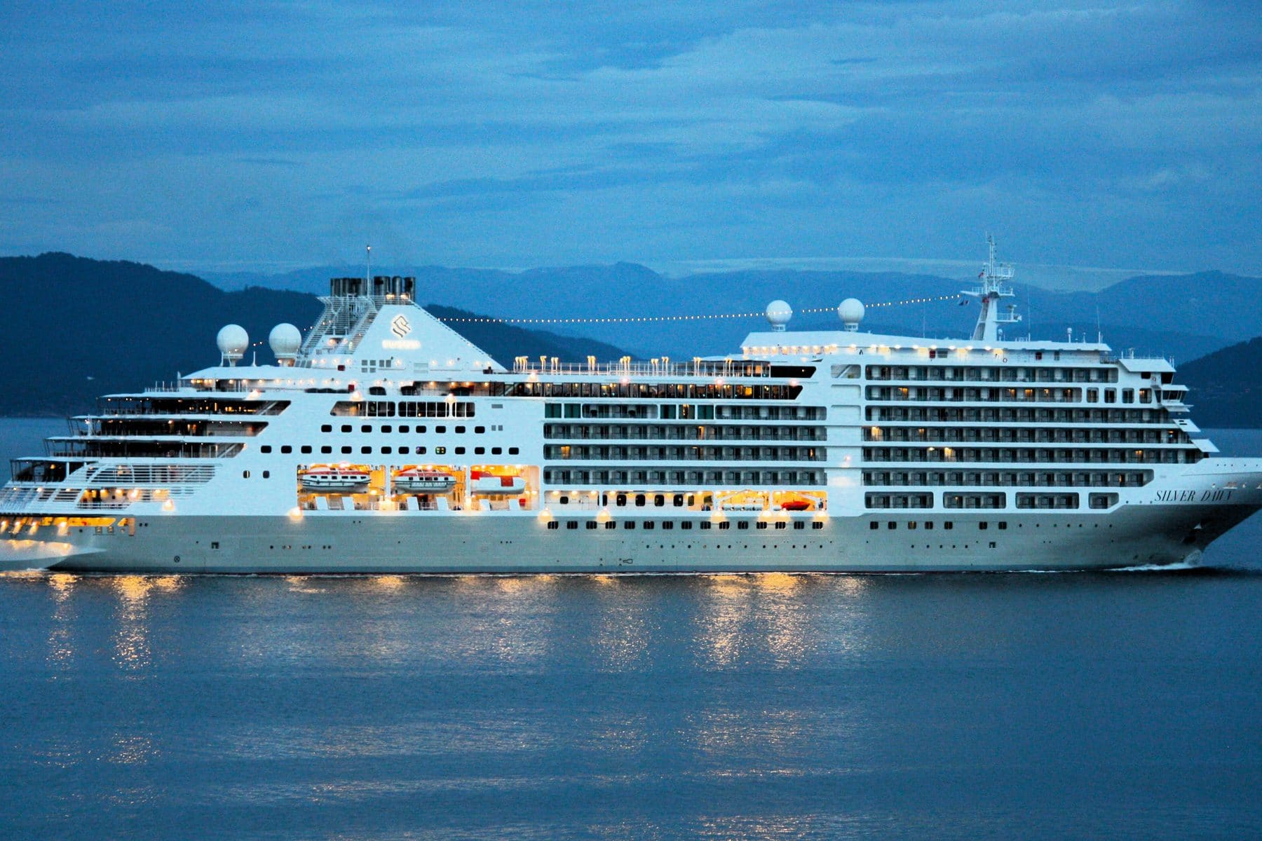 Silver Dawn at dusk, lights on along the decks and a mountain shore behind